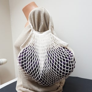 May include: A white string mesh bag with a purple fabric item inside. The bag is being held by a person wearing a beige sweater.
