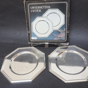Può includere: Due piatti da portata ottagonali in argento con un bordo rialzato. I piatti sono in una scatola che dice "Untersetzer 2 Stuck".