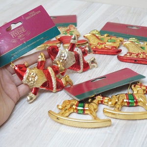 Puede incluir: Un conjunto de seis adornos para árbol de Navidad de color dorado y rojo. Los adornos incluyen dos Papá Noeles, dos trineos y dos caballos de balancín. Los adornos están empaquetados en una tarjeta roja y verde con las palabras "Christmas Finesse" impresas en ella.