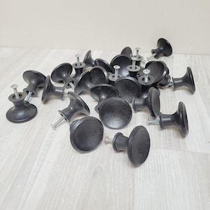 May include: A collection of black cabinet knobs with silver screws. The knobs have a rounded, mushroom-like shape and are scattered across a light-coloured wooden surface. The knobs are made of a matte material.