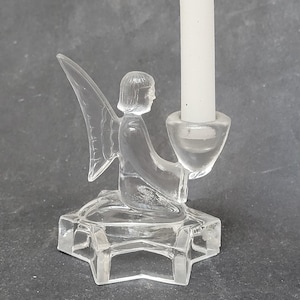 Könnte beinhalten: Transparenter Glas-Engels-Kerzenhalter mit einer weißen Kerze. Der Engel kniet auf einem sternförmigen Sockel und hält eine Kerzenschale. Die Flügel sind ausgebreitet. Ein dekorativer Artikel.