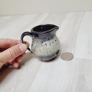 May include: A small, white and blue ceramic creamer with a speckled glaze. The creamer has a rounded body and a small spout.