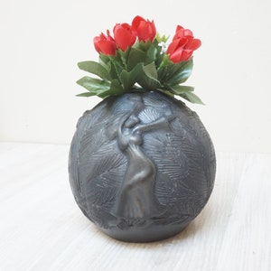 Könnte beinhalten: Eine schwarze Keramikvase mit einem Relief einer tanzenden Frau. Die Vase ist rund und hat eine strukturierte Oberfläche. Sie ist mit roten Blumen gefüllt.