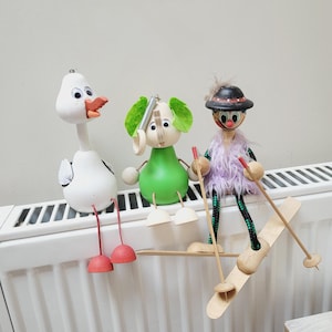 May include: Three wooden figurines, a white duck, a green elephant, and a purple-clad skier, are perched on a white radiator. The skier is holding two wooden ski poles and is on a wooden ski.