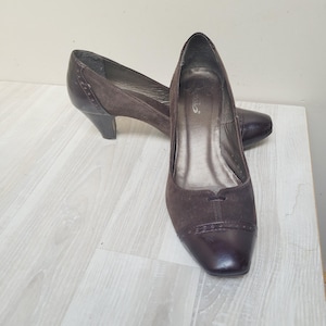 May include: A pair of dark brown pumps featuring a suede upper with a smooth leather cap toe and heel counter. The shoes have a low kitten heel, a square toe shape, and a decorative perforated trim detail along the seam of the toe cap.