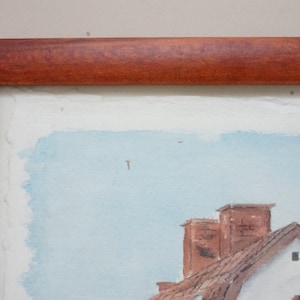 May include: Watercolor painting of a village scene, framed in a dark wood. The artwork depicts buildings with red-tiled roofs and chimneys against a light blue sky. The painting has a rustic, traditional style.