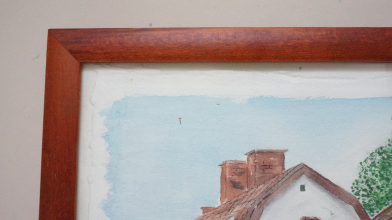 May include: Watercolor painting of a village scene, framed in a dark wood. The artwork depicts buildings with red-tiled roofs and chimneys against a light blue sky. The painting has a rustic, traditional style.