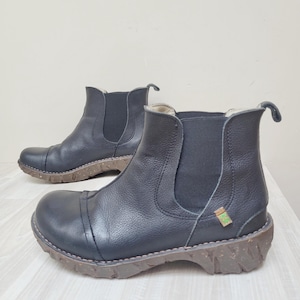 May include: A pair of black leather ankle boots featuring a rounded toe, elastic side panels, and a thick, textured brown rubber sole. Each boot includes a small green tag on the side and a pull tab at the heel for easy access and removal.