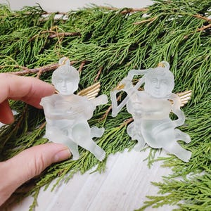 Könnte beinhalten: Zwei Engel-Ornamente aus mattiertem Glas mit goldfarbenen Flügeln und Aufhängeschlaufen. Ein Engel spielt ein Saiteninstrument, der andere hält einen Bogen und einen Pfeil. Die Ornamente sind vor einem Hintergrund aus grünen Tannenzweigen angeordnet.