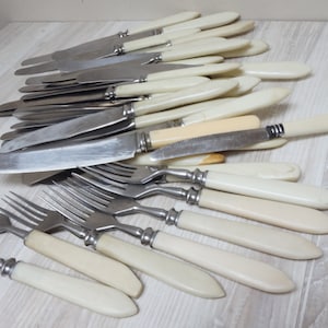 Puede incluir: Un conjunto de cubiertos antiguos, que incluye cuchillos y tenedores. Los cuchillos tienen hojas de acero inoxidable y mangos de color crema. Los tenedores tienen cuatro púas y mangos a juego. Los cubiertos están dispuestos sobre una superficie de madera clara.