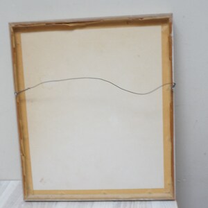 May include: The back of a framed picture with a light brown wooden frame and a white mat. A thin wire is attached to the frame for hanging.