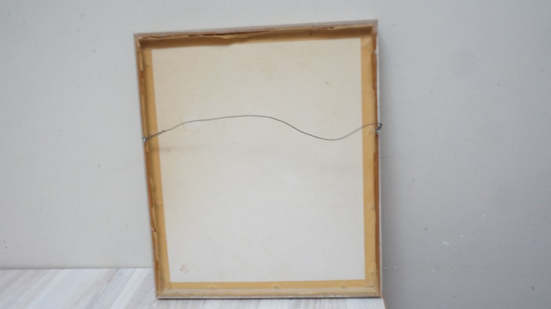May include: The back of a framed picture with a light brown wooden frame and a white mat. A thin wire is attached to the frame for hanging.