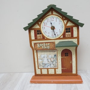 Könnte beinhalten: Eine Holz-Uhr in Form eines Friseursalons. Das Zifferblatt ist weiß mit schwarzen Zahlen und Zeigern. Das Gebäude hat ein grünes Dach und ein Schild mit der Aufschrift "Barber Shop". Die Uhr steht auf einem Holzsockel.