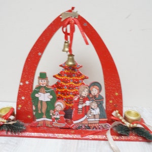 K&ouml;nnte beinhalten: Roter Weihnachtsbogen aus Holz mit einer gemalten Szene einer Familie, die Weihnachtslieder um einen Weihnachtsbaum singt. Der Bogen hat zwei Kerzenhalter auf jeder Seite und eine Glocke, die oben h&auml;ngt.