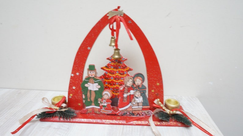 K&ouml;nnte beinhalten: Roter Weihnachtsbogen aus Holz mit einer gemalten Szene einer Familie, die Weihnachtslieder um einen Weihnachtsbaum singt. Der Bogen hat zwei Kerzenhalter auf jeder Seite und eine Glocke, die oben h&auml;ngt.