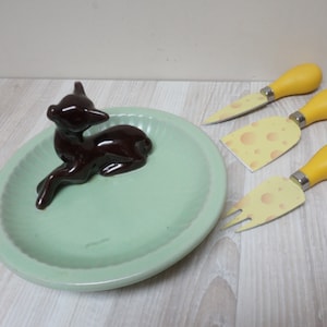 Peut inclure: Une assiette en céramique verte avec une figurine de cerf brune. Trois couteaux à fromage jaunes avec un motif de fromage sont sur une surface en bois.