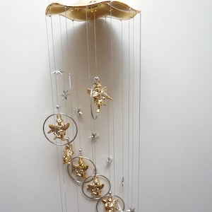 Puede incluir: Un carillón de viento dorado con una parte superior en forma de nube y múltiples colgantes. Los colgantes incluyen pequeñas estrellas y ángeles con alas.