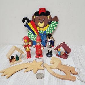 May include: A collection of vintage wooden Christmas ornaments, including a teddy bear, a star, a rocking horse, a nativity scene, and two figures in red and blue uniforms.