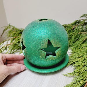May include: A green, glitter-covered sphere-shaped candle holder with star cutouts, sitting on a matching round base. The candle holder is surrounded by green foliage, creating a festive holiday theme.