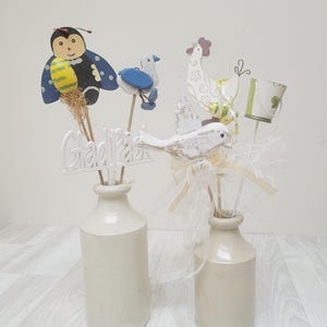 May include: Two beige ceramic bottles holding decorative wooden sticks featuring a ladybug, a blue bird, a white chicken, a white bird, and a small bucket. A white cutout sign with the text Glad Påsk is attached to one of the wooden sticks.