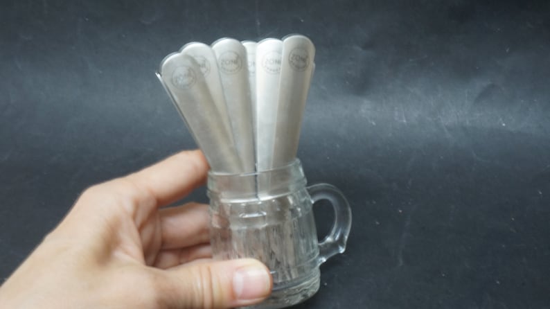 K&ouml;nnte beinhalten: Ein Set aus sechs silbernen L&ouml;ffeln mit einem Logo am Griff, angeordnet in einer gl&auml;sernen Tasse mit Henkel.