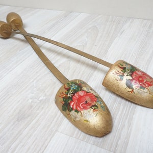 May include: A pair of antique wooden shoe trees with floral designs painted on them. The shoe trees are gold in color and have long, thin metal rods connecting them.