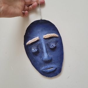 Könnte beinhalten: Eine blaue Keramikmaske mit einem traurigen Ausdruck. Die Maske hat weiße Augenbrauen, blaue Augen und einen blauen Mund. Sie hängt an einer Schnur.