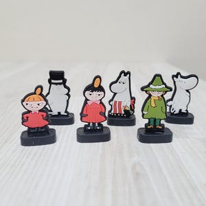 May include: A collection of seven cartoon character figurines. The figurines are in various colors, including red, green, and black. Each figure is mounted on a small, rectangular black base. The characters appear to be from a children's cartoon.