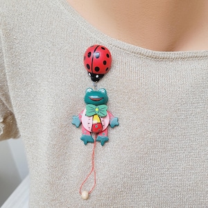 May include: A wooden frog brooch with a red bow tie and a ladybug hanging from its feet. The frog is green, pink, and yellow. The ladybug is red with black spots.