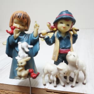 Op de afbeelding: Twee beeldjes die een herder en een engel uitbeelden. De engel draagt een blauwe jurk en heeft een stapel konijnen. De herder heeft een hoed, draagt een staf en wordt vergezeld door drie lammeren. De beeldjes zijn in verschillende kleuren geschilderd.