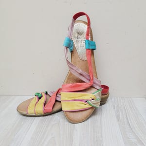 Peut inclure: Une paire de sandales en cuir multicolores avec une bride arrière, une semelle plate et un design noué sur le devant. Les lanières sont de couleur rouge, jaune, rose et bleu clair. La semelle intérieure est beige avec la marque Marila imprimée.