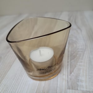 May include: A clear glass candle holder with a triangular shape and a brown tint. A white tea light candle is inside the holder. The holder has the text "Florian Platini" and "Design Studio Johannesburg" printed on the bottom.