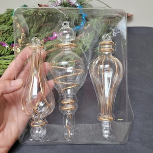 May include: Three clear glass Christmas ornaments with gold accents and hanging loops are displayed in a clear plastic package. The ornaments have different shapes, including a teardrop, a bulb, and a slender design. The gold detailing adds a touch of elegance to the holiday decorations.