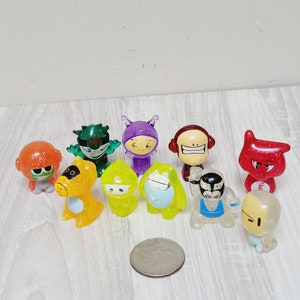 Puede incluir: Una colección de diez pequeñas figuritas de plástico de colores con purpurina. Las figuritas son personajes variados con diferentes expresiones faciales y poses. Las figuritas están sobre una superficie de madera con una moneda de plata.