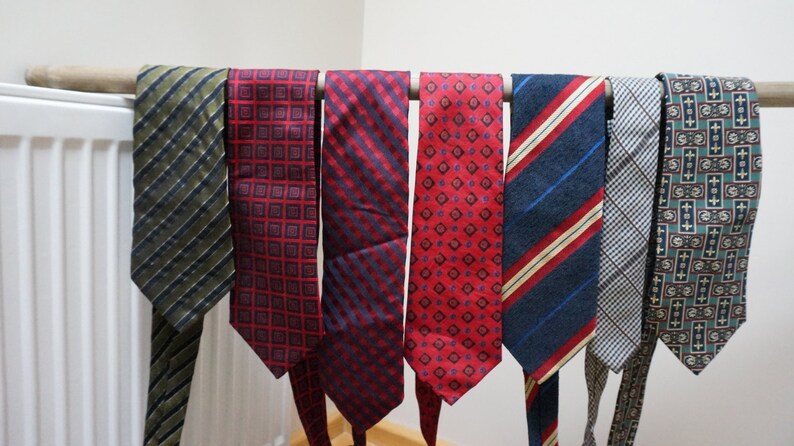 Puede incluir: Ocho corbatas de diferentes patrones colgando de una barra de madera. Las corbatas son de varios colores, incluyendo rojo, azul, verde y negro.