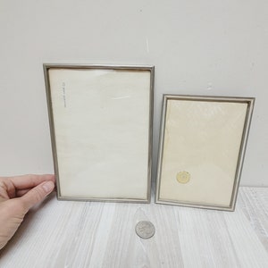 Könnte beinhalten: Zwei silberne Bilderrahmen mit Glasfronten. Der größere Rahmen befindet sich links und der kleinere Rahmen rechts. Der größere Rahmen hat eine kleine Inschrift oben links, die "10 Jahre Garantie" lautet.