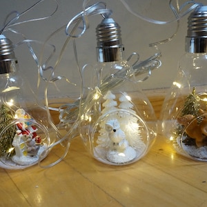 Puede incluir: Tres bombillas de vidrio transparente con una cadena de luces en su interior. Cada bombilla contiene una escena invernal en miniatura con una pequeña figura, incluyendo un muñeco de nieve, un búho y un ciervo.