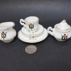 Può includere: Un set da tè in miniatura in porcellana bianca con dettagli dorati. Il set comprende una teiera, un creamer, uno zuccheriera, una tazza e un piattino. I pezzi sono decorati con una ghirlanda verde e uno stemma rosso e oro con la scritta "Harrods" stampata sul piattino.
