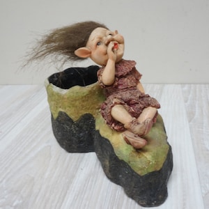 May include: A whimsical figurine of a gnome with brown hair sitting on a green and black boot-shaped planter. The gnome is wearing a pink dress and has a playful expression.