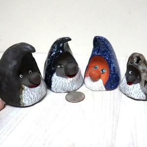 Puede incluir: Cuatro gnomos de cerámica con sombreros y caras de diferentes colores. Los gnomos llevan todos barbas blancas y tienen ojos negros. Los gnomos están sentados sobre una superficie de madera blanca.