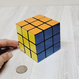 May include: A black, yellow, and blue 3x3 Rubik's Cube puzzle. The cube is sitting on a light wood surface.