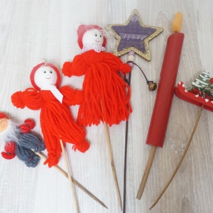 Op de afbeelding: Een verzameling kerstversieringen op houten stokken. Inclusief rode garendpoppen, een paarse ster, een rode kaars en een slee met een kerstboom en een beer. De items zijn gerangschikt op een licht houten oppervlak.