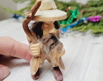 May include: A hand-carved wooden figurine of an elderly man wearing a hat and holding a walking stick. The figure is detailed with carved facial features and clothing, and is made of light and dark brown wood. The figurine is standing on a light-colored surface.