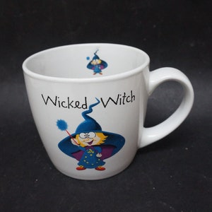 May include: White ceramic mug with a cartoon witch design. The mug features the words "Wicked Witch" and an illustration of a smiling witch in a blue hat and cape, holding a wand. The mug has a curved shape and a large handle.