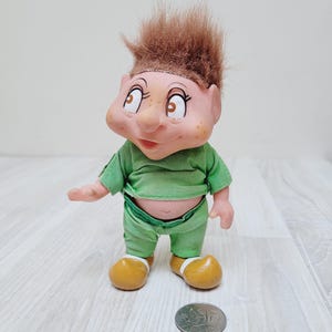 May include: A vintage troll doll with a painted face, freckles, and brown hair. The doll wears a green outfit and yellow shoes. A silver coin is next to the doll.