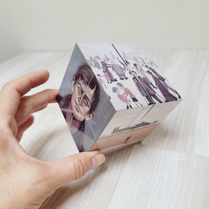 Pode incluir: Um quebra-cabeça em forma de cubo com ilustrações em cada lado. Um lado mostra um retrato de um homem com cabelo escuro e olhos intensos. Outros lados mostram cenas de pessoas com roupas vintage. O quebra-cabeça é segurado por uma mão.