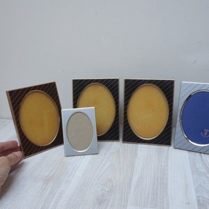 Puede incluir: Cinco marcos de fotos ovalados con ribete dorado. Los marcos están dispuestos en una fila sobre una superficie de madera clara. Los marcos son de diferentes tamaños y tienen fondos de colores diferentes. Un marco tiene un fondo marrón, dos tienen un fondo dorado y uno tiene un fondo azul.