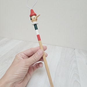 May include: A wooden pencil featuring a carved Pinocchio character topper with a red hat, a long nose, and painted facial features. The pencil body is painted with horizontal bands of dark green, white, and red, transitioning to a natural wood finish.