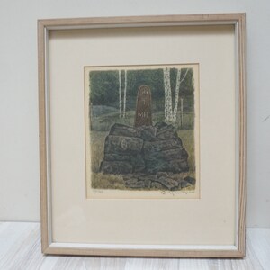 May include: A framed print of a wood carving depicting a figure standing on a pile of rocks in a forest setting. The carving is brown and the background is green and white.