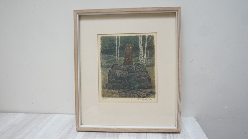 May include: A framed print of a wood carving depicting a figure standing on a pile of rocks in a forest setting. The carving is brown and the background is green and white.